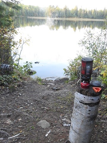 a red self-contained cooking stove set up on a birch log on the shore of Chickenbone Lake at West Chickenbone campground