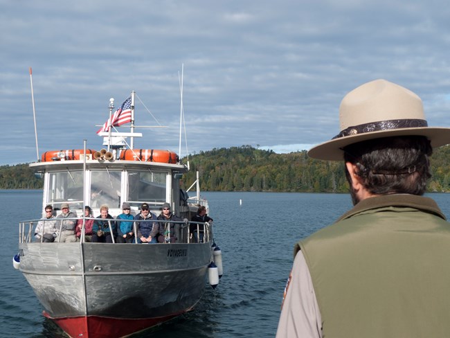 A steel hulled boat approaches a ranger on a dock. People are sitting on the bow.