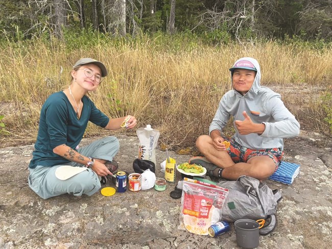 Two people sit on a rock surrounded by food.