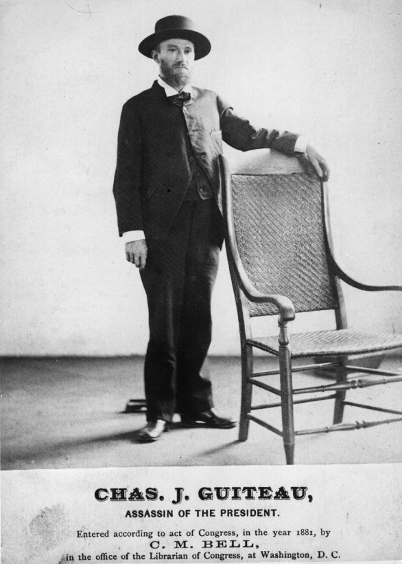 man in a suit standing to the left of a wicker chair with his arm on the back of it