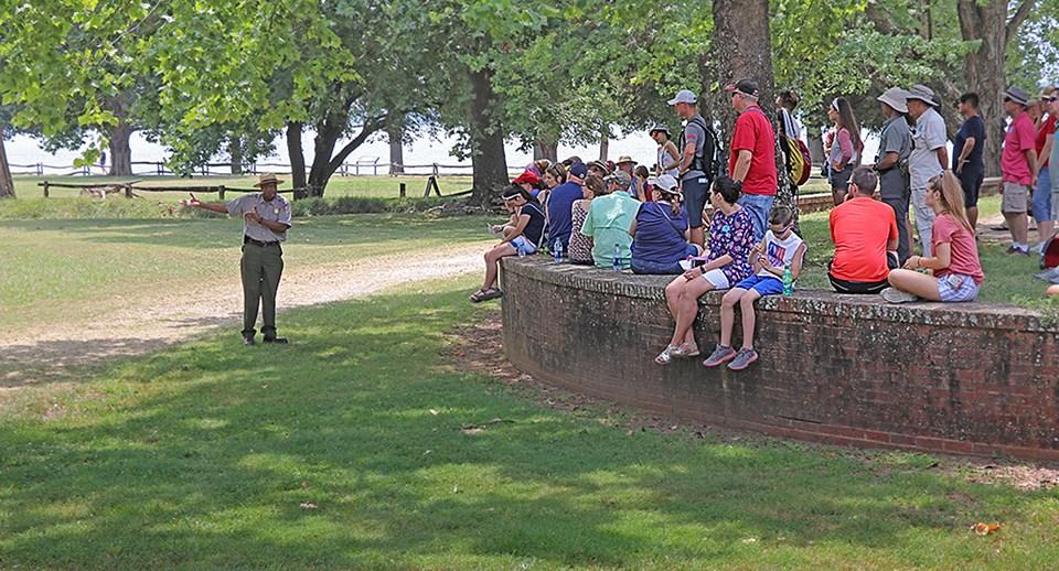 A park ranger talks to a group of visitors. The group sits along a curved brick wall in a grove of trees.