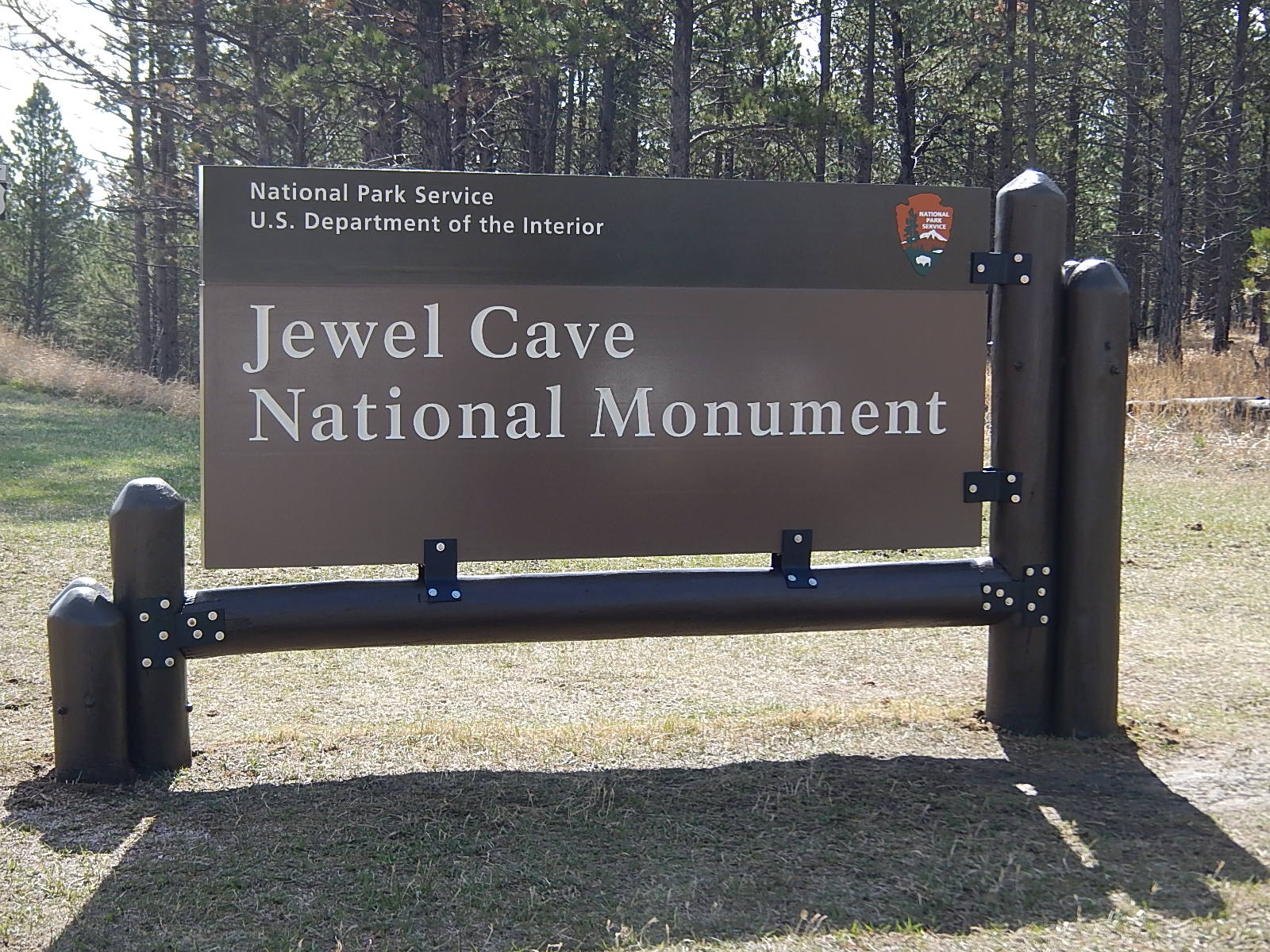 A large sign reads "Jewel Cave National Monument" surrounded by short grass with a forest background.