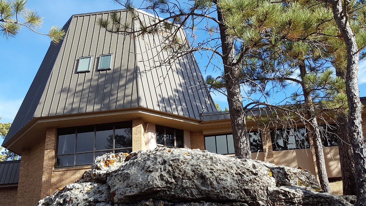 Jewel Cave Visitor Center A brick building with a tall brown roof surrounded by evergreen trees under a blue sky