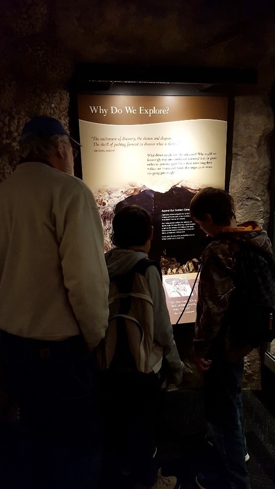 One adult and two youth standing in a dimly lit exhibit area viewing an interpretive sign.