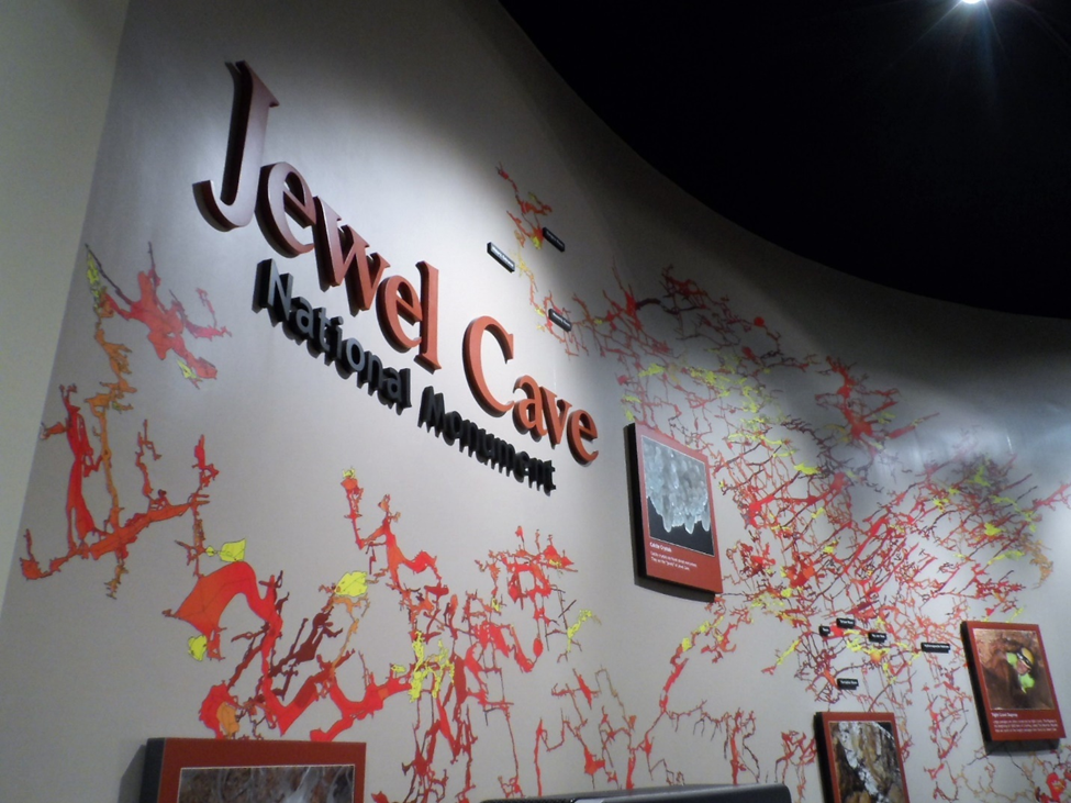A sign reading Jewel Cave National Monument in front of a multicolored cave map