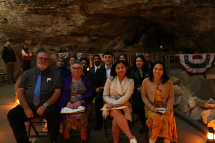 Twelve smiling people sit in chairs surrounded by rock walls and red, white, and blue flags.