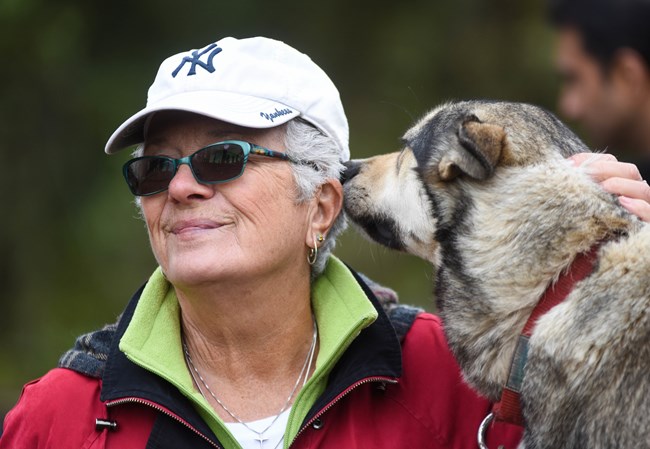A woman wearing a white ballcap is being snuggled by a large dog.