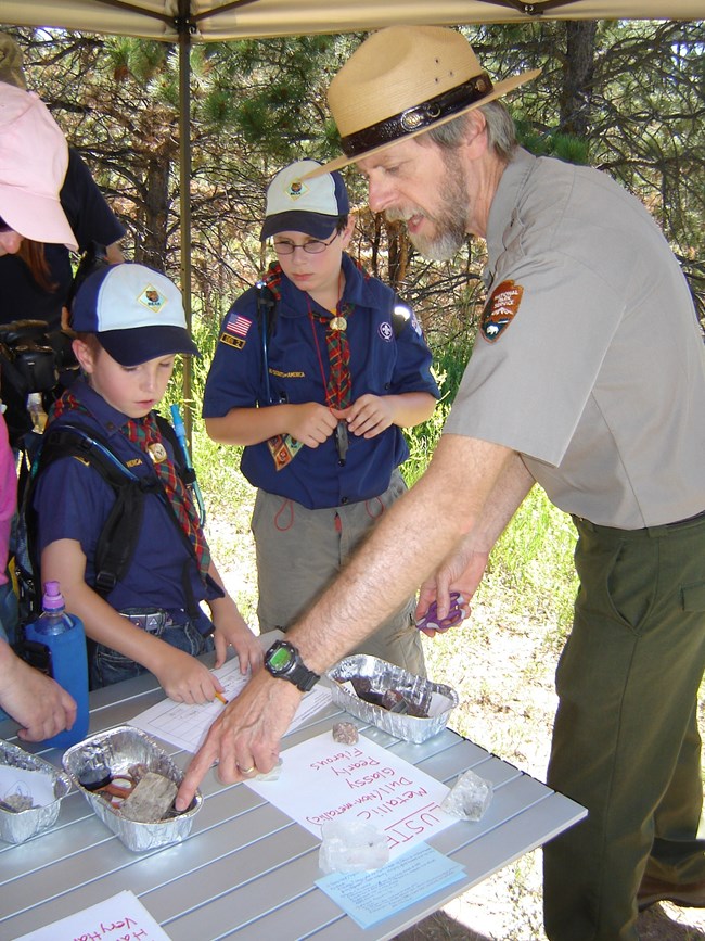 A park ranger shows a group of Cub Scouts and their chaperones some rock samples on a folding table.