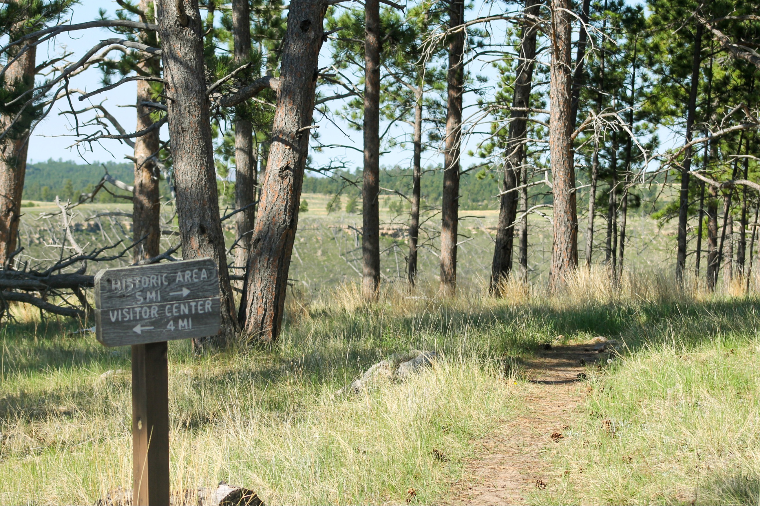 A brown trail sign with distances to the Historic Area and Visitor Center, showing a worn path in the background.