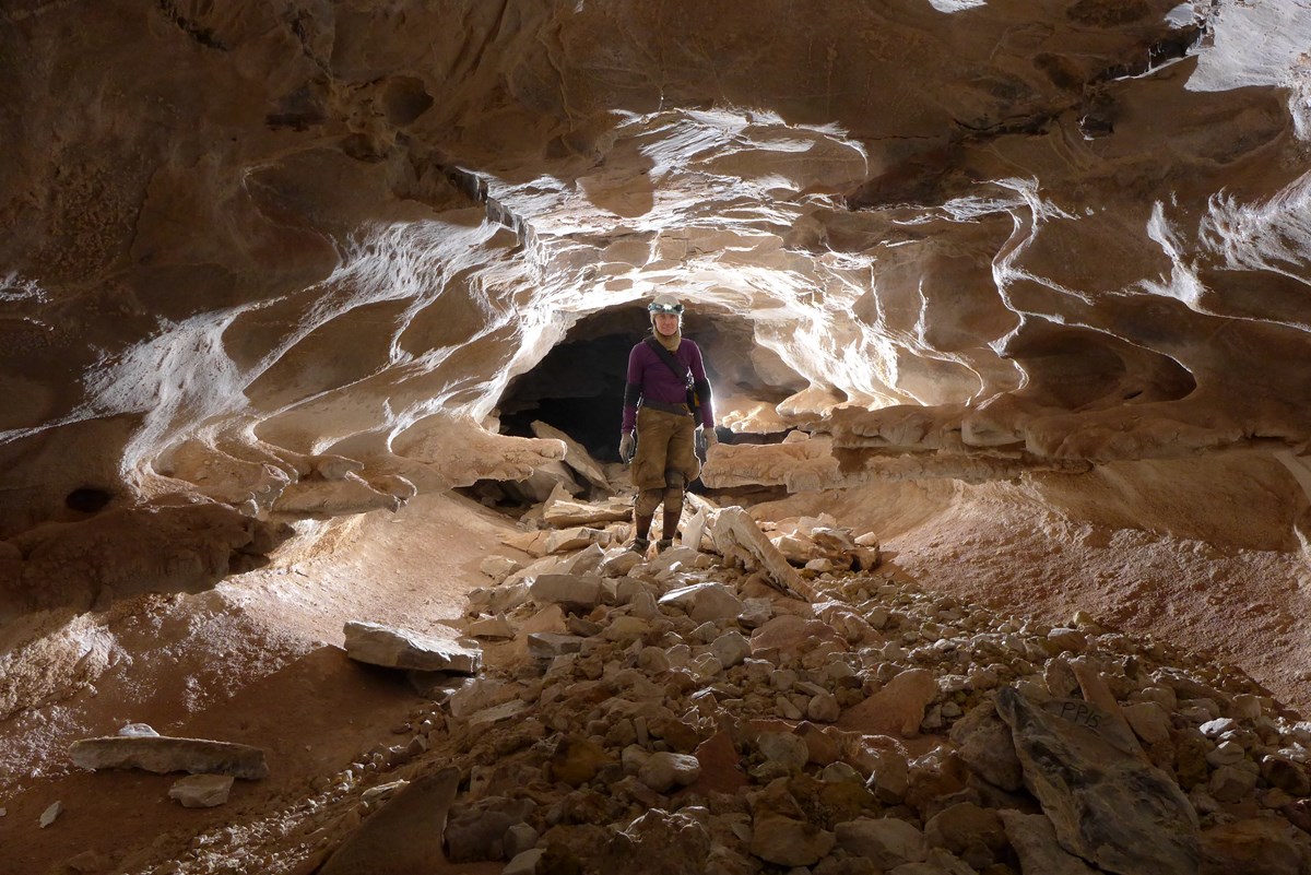 A cave explorer stands in passageway, with backlighting behind the picture.