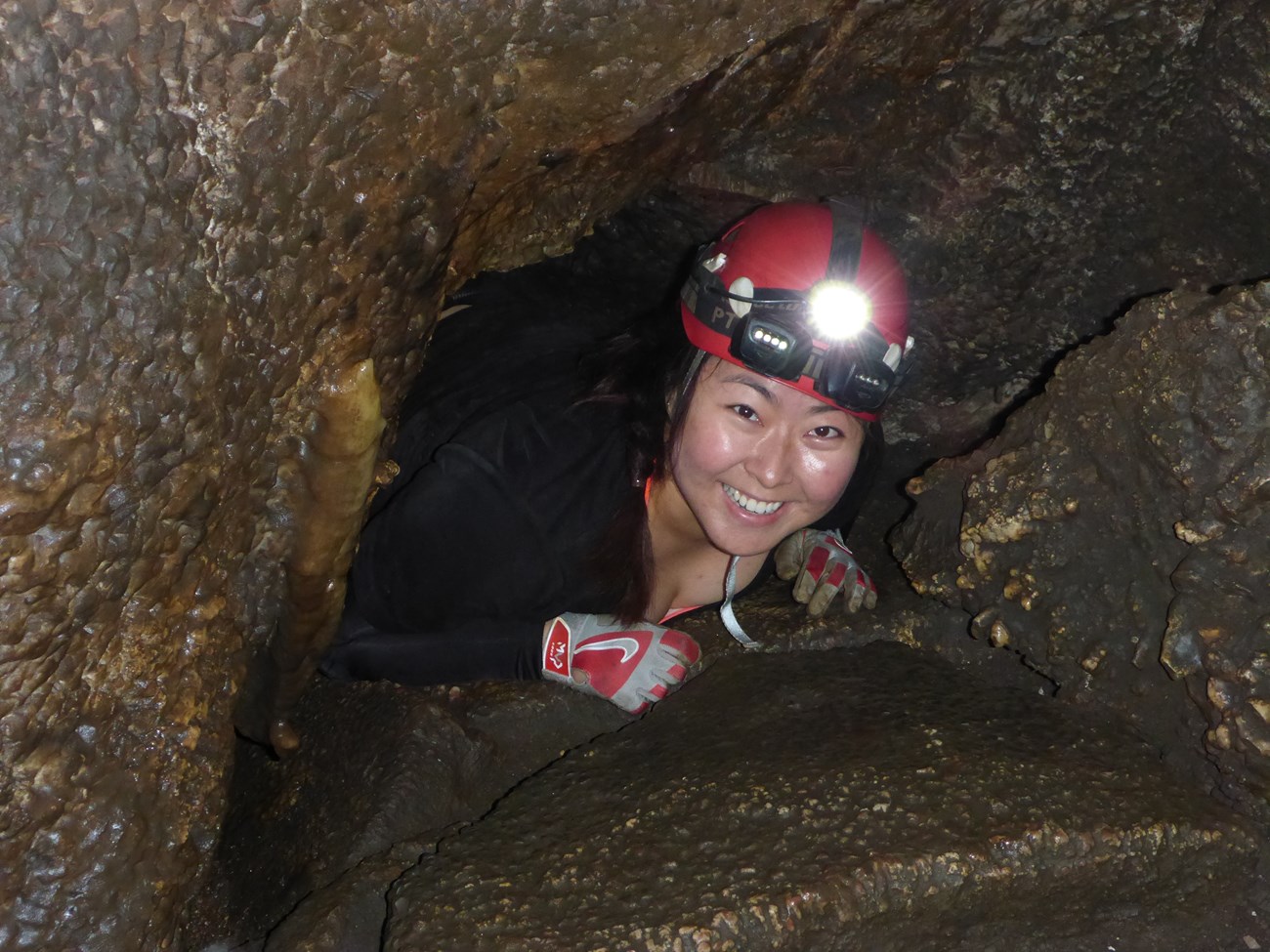 A caver wearing a helmet and headlamp squeezes through a tight opening.