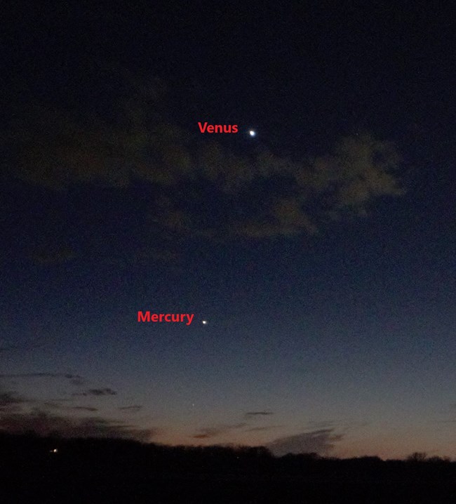 Dark blue sky with orange twilight and a few white clouds. A line of trees marks the horizon. White dots mark the planets Mercury and Venus.