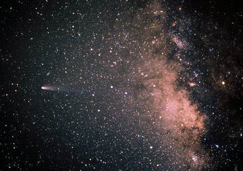 Stars form white dots on the black field of the sky.  Pink clouds form the Milky Way, with a comet showing a short tail pointing to the right in the view.