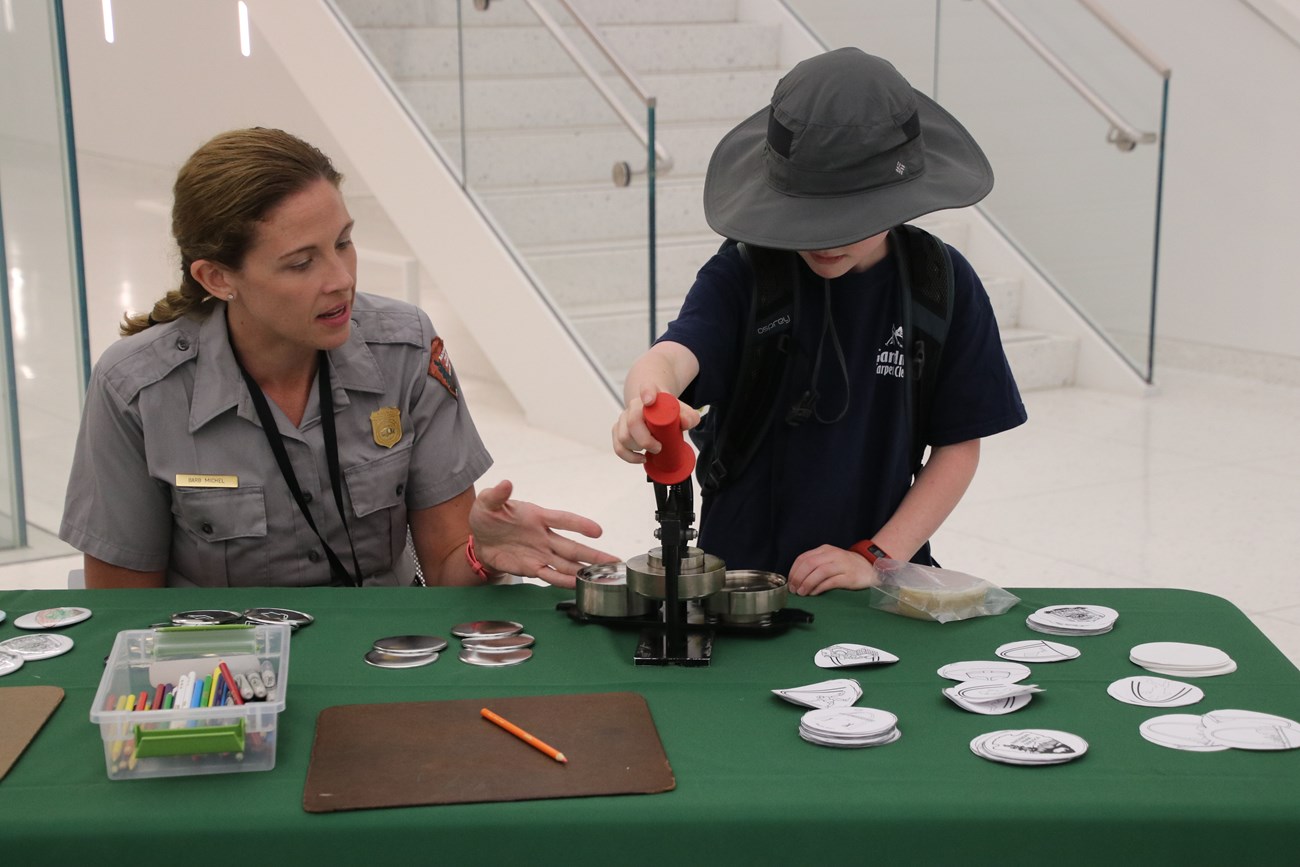 Park Ranger sits with student at table as the student uses a button-making device.