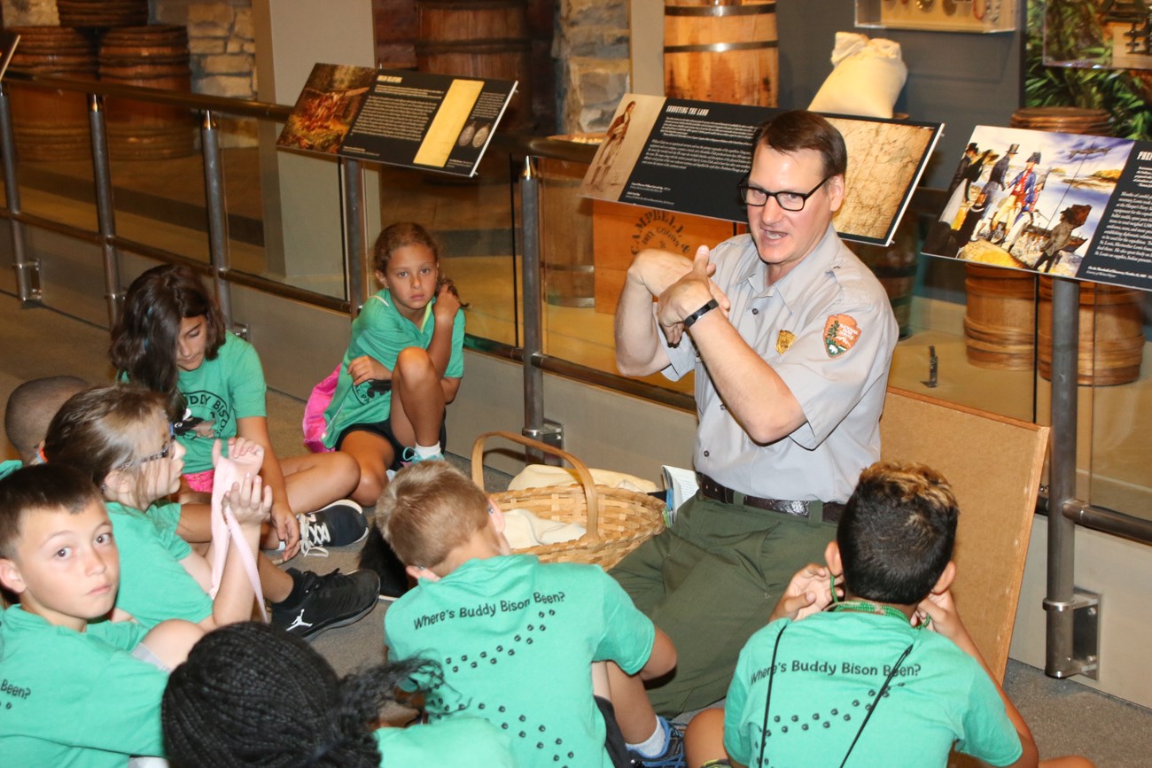 Park Ranger sits on the floor with students and leads a program with exhibits in the background.