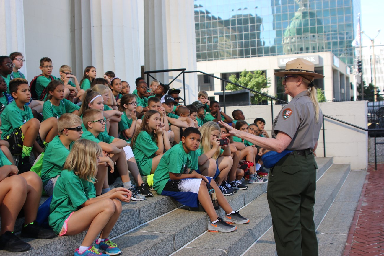 Park Ranger in green and gray uniform addressing students sitting on courthouse steps.