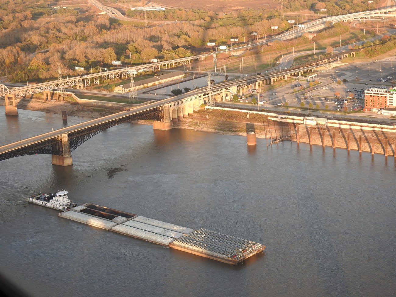 A towboat pushing several barge cars passes under a bridge. A shadow of an arch is on the opposite riverbank.