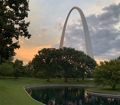 South pond in the foreground with the Gateway Arch in the background with an orange and blue sky. Green grass and trees surround the Arch and pond.