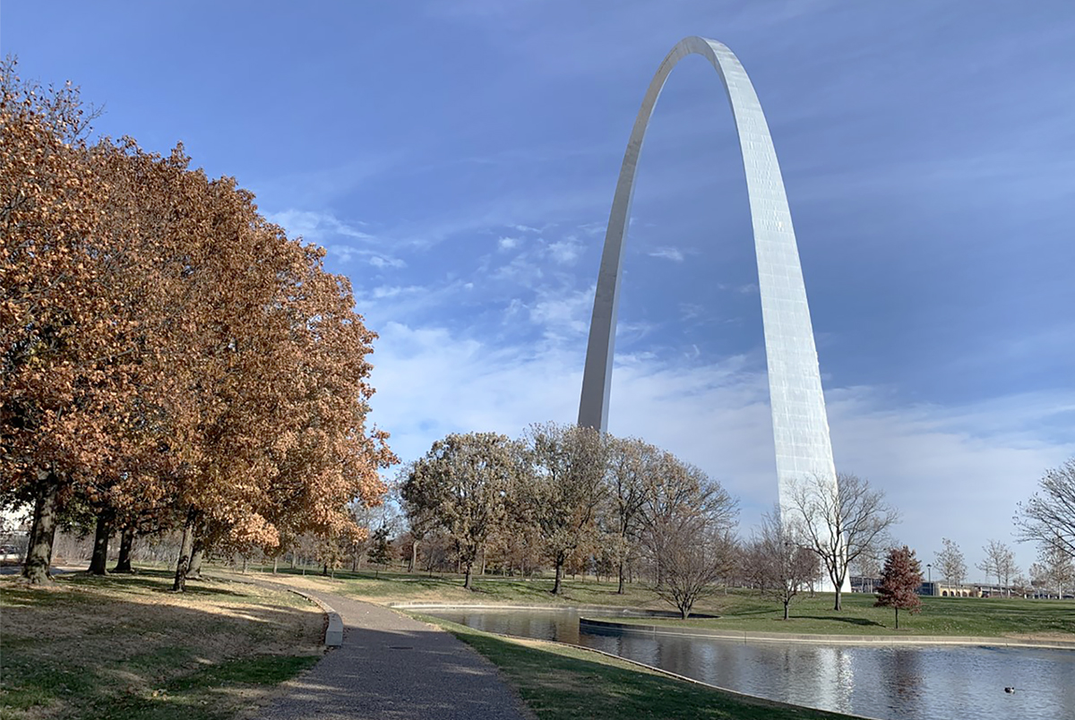 The Gateway Arch stands off to the right. On the left are large trees with brown leaves. A sidewalk curves around the trees and disappears into the distance. To the right of the sidewalk is the artificial south pond which reflects part of an Arch leg and