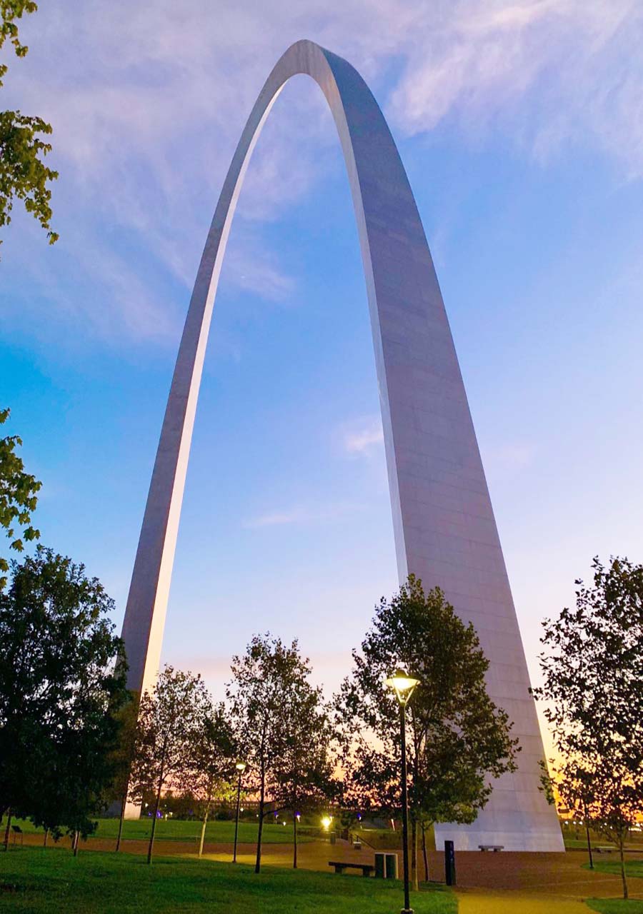 Gateway Arch at sunrise. A yellow glow behind the south leg of the Arch indicates the sunrise. White puffy clouds and a blue sky are behind the Arch.