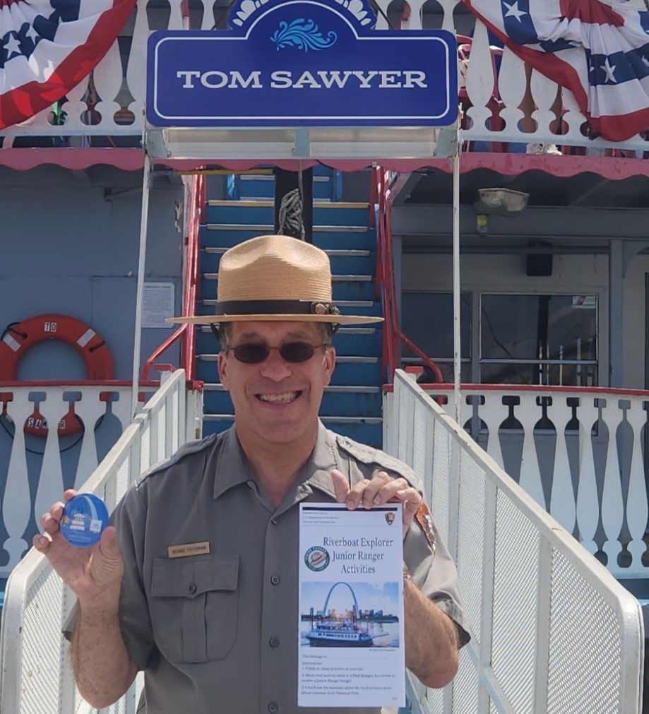Ranger and Riverboat Male park ranger standing in front of the Tom Sawyer Riverboat. He is holding the Junior Ranger Riverboat booklet and the button you get when you complete the booklet.