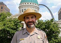 A man smiles at the camera. He's wearing a uniform with a flat hat. A white building with a green dome and the Gateway Arch are behind him.