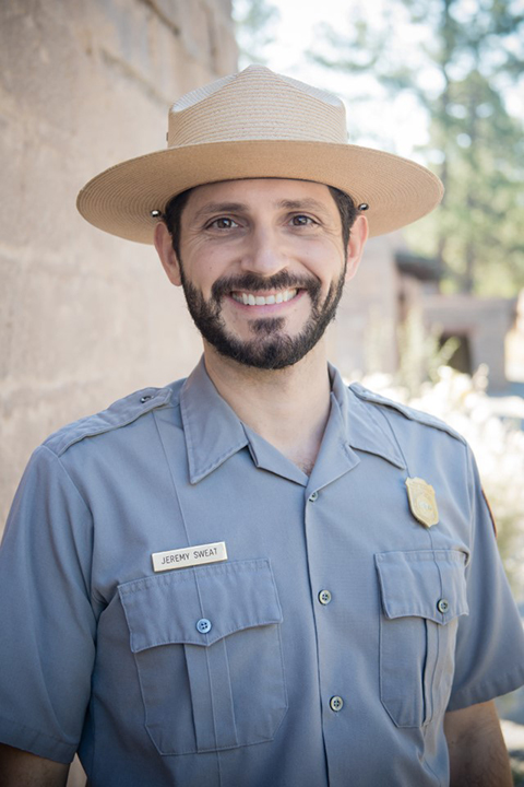 Portrait of Jeremy Sweat in uniform from the chest up with a building and trees behind.