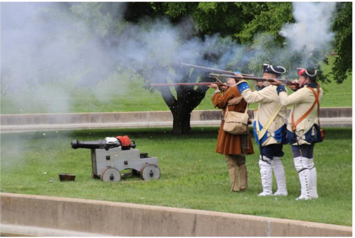 Three men in 18th century clothing fire muskets next to a cannon