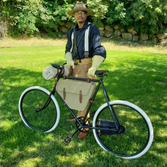A man wearing late-1800s clothing stands next to a historic bicycle