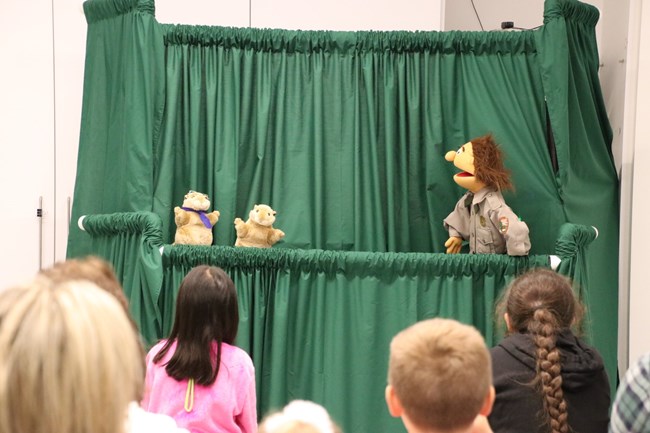 The backs of children's heads, facing several puppets on a stage.
