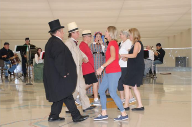 Lines of dancers, some in 19th century clothing and some in modern clothing, stand in two lines.