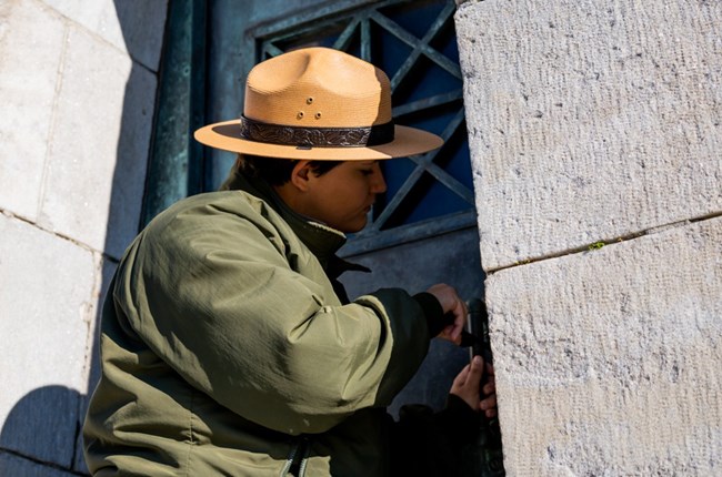 A park ranger wearing a green jacket and straw flat hat unlocks the metal door to the Chalmette Monument