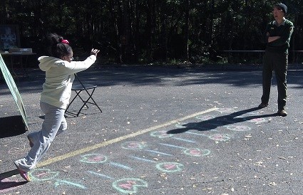 A female ranger watches as a young girl leaps across chalk lines on pavement