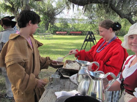 Image of young women in 1815 dress serving lunch to young men in period militia clothes