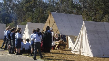 A group of school children in blue uniforms speak to history reenactors. Tan triangle tents are set up on grass.