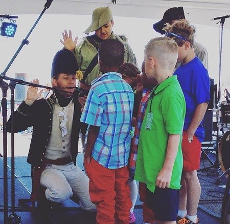A reenactor in War of 1812 attire with right hand raised, kneeling with five boys and girls being sworn in as junior rangers