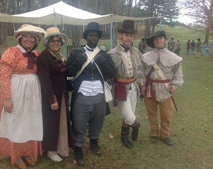 Young men and women in living history attire of the War of 1812 pose in front of a tent and a cannon