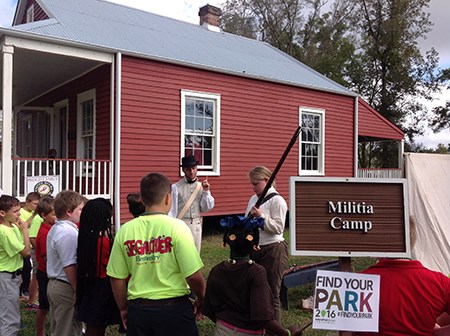 A ranger and a volunteer in living history attire of the War of 1812 display a musket to a group of elementary school student.