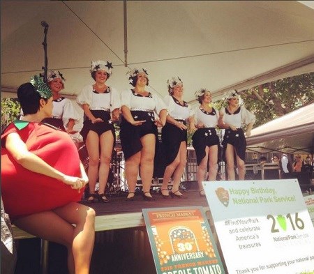 A line of six chorus girls dance on a stage and a woman in a tomato costume sits at the edge of the stage