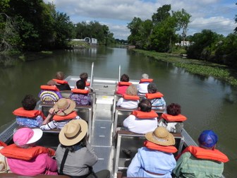 Image is people in an open boat heading up a bayou