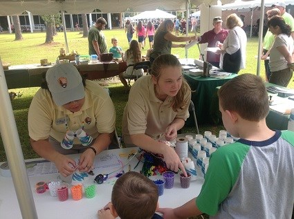 Two Centennial Volunteer Ambassadors assist a young park visitor to construct a craft project with colored beads