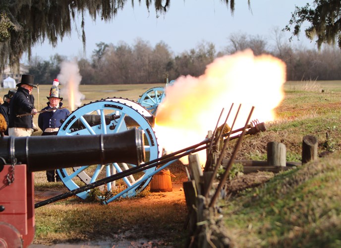 Image of men in 1815 uniforms firing a cannon; fire roars from the cannon's mouth