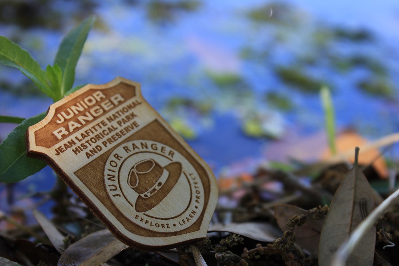 A wooden badge that says "Junior Ranger Jean Lafitte National Historical Park and Preserve" with a hat logo. Water is in the background.