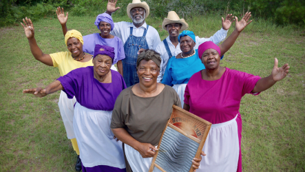 Press Release__Gullah Image A group of Gullah GeeChee people smiling together with arms spread out. One person in the front holds a washboard up.