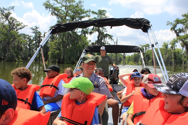 A park ranger and kids sit in a metla boat wearing orange life jackets smiling in a swamp.