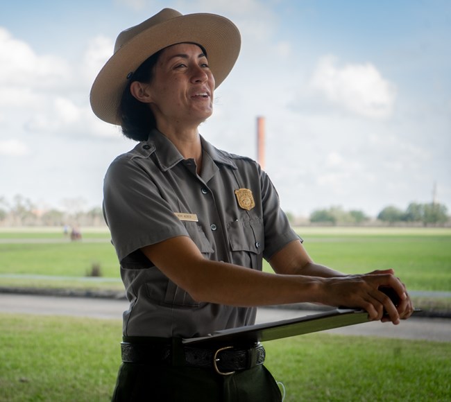 A park ranger in a gray uniform shirt and flat hat smiles and holds a clipboard.
