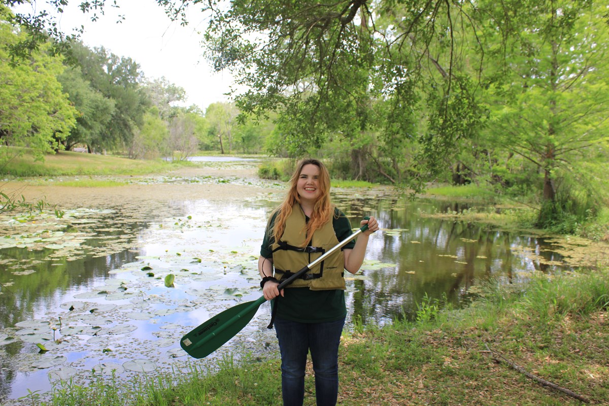 A girl stands next to a bayou. She holds a canoe oar in her hands, and she is wearing a life jacket.