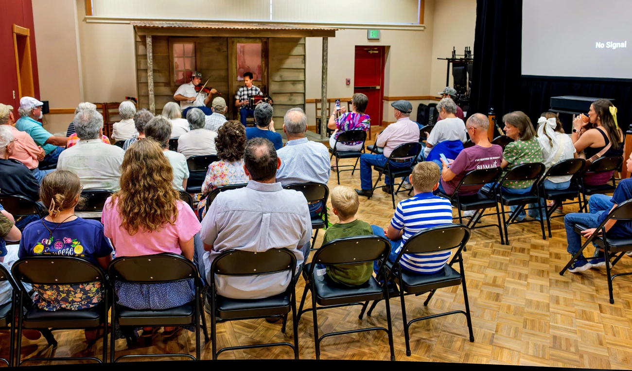 Experience culture live at the Prairie Acadian Cultural Center An audience of people sitting and listening to two musicians perform on a front porch stage.