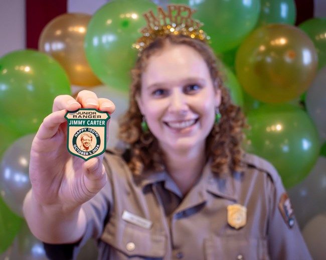 A park ranger holds a special edition Junior Ranger badge celebrating Jimmy Carter's 100th birthday, with colorful balloons floating in the background.