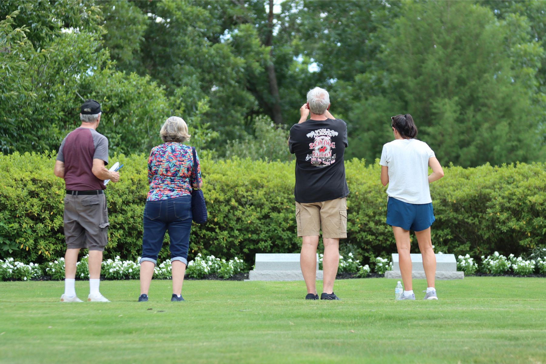 Visitors pay their respects at the gravesite of President Jimmy Carter and First Lady Rosalynn Carter. Four visitors facing the gravesite of Jimmy and Rosalynn Carter, with lush green hedges and trees in the background.